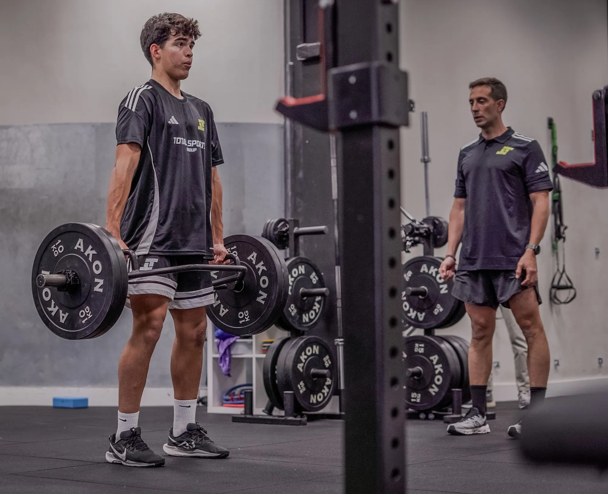 A player lifting a loaded barbell in the gym while a coach supervises